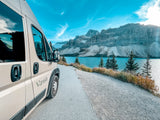 Scenic rv rental adventure! Beige campervan overlooks a vivid blue lake, rugged mountains, and a bright, cloudy sky.