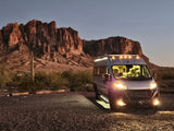 An RV rental camper van, lit up at dusk, parked on a gravel lot with majestic desert mountains in the background.