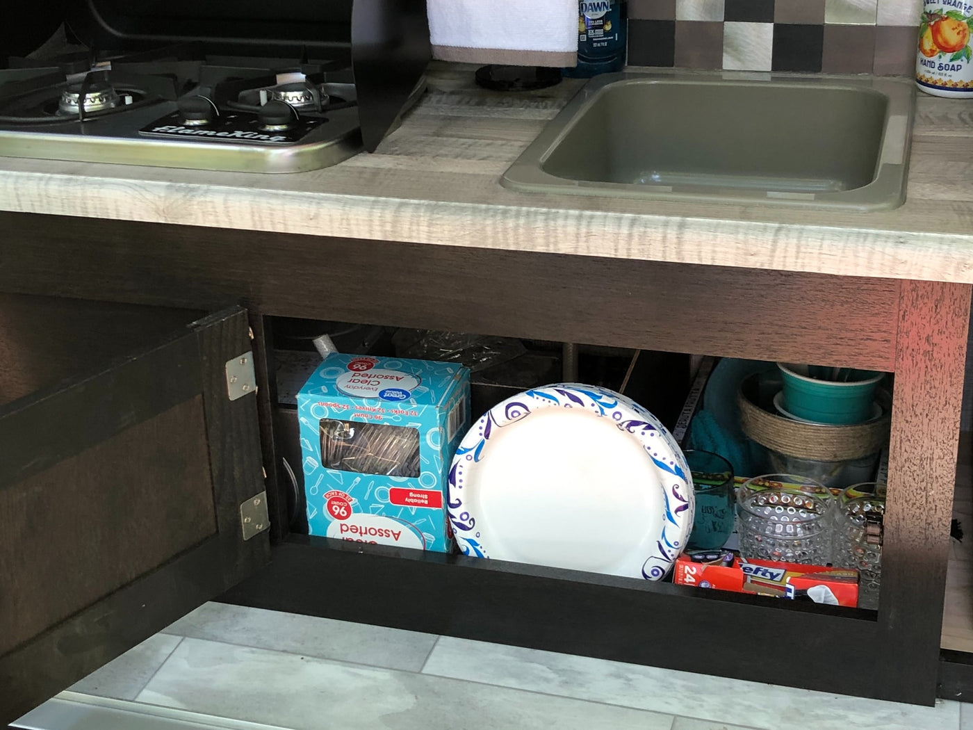 RV rental kitchen: Open cabinet stocked with disposable plates, cutlery, glasses. Sink, stove, & cleaning supplies on counter. — photo 20