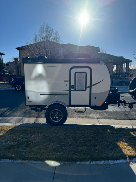 A modern, light-colored travel trailer with a Thule awning, perfect for an RV rental, parked on a sunny street.