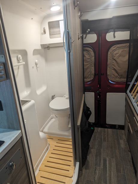 RV rental: A compact camper van wet bath with toilet, sink, shower, and a wooden floor mat. Rear doors visible.