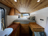 Cozy RV rental interior: wood-paneled ceiling, white walls, comfortable bed, compact kitchen sink. Mountain views outside.