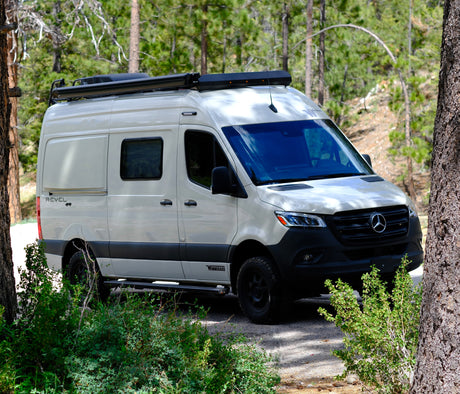 White Mercedes-Benz Sprinter Winnebago Revel 4x4 camper van in a forest, perfect for an RV rental adventure.