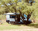 A white RV rental campervan with an extended awning and picnic table sits shaded by trees at a grassy campsite.