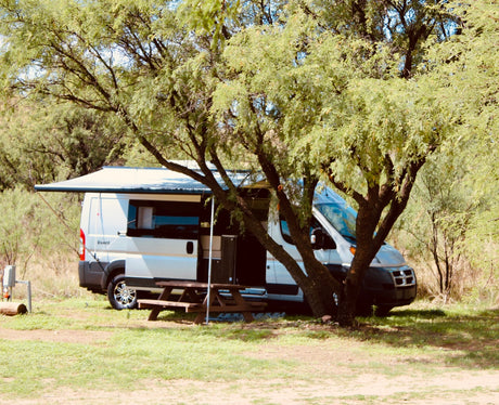A white RV rental campervan with an extended awning and picnic table sits shaded by trees at a grassy campsite.