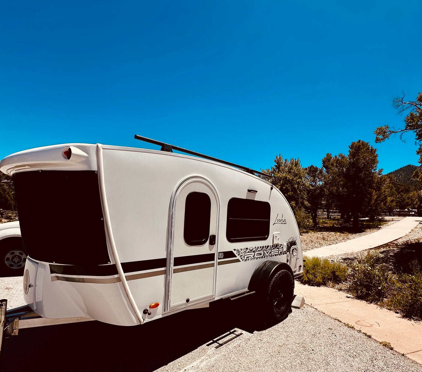 A white Luna Rover teardrop RV rental parked in a sunny camp spot with a clear blue sky and pine trees.