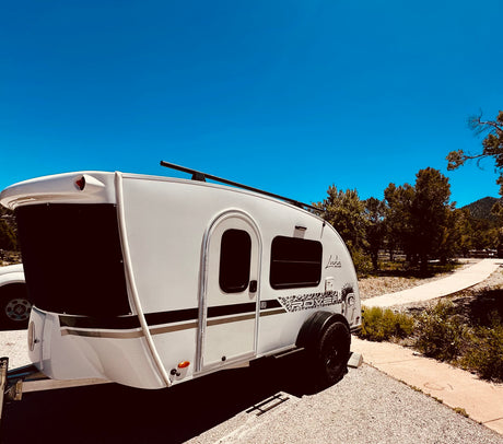 A white Luna Rover teardrop RV rental parked in a sunny camp spot with a clear blue sky and pine trees.