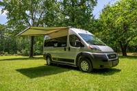 Enjoy the outdoors with this RV rental! A silver campervan, with a large awning extended for shade, parked on green grass. — photo 7