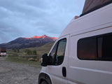 White camper van (rv rental) by a tranquil lake, mountains bathed in alpenglow. A perfect nature escape awaits.