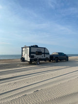 An RV rental setup: a truck and travel trailer parked on a wide sandy beach by the ocean, tire tracks visible.