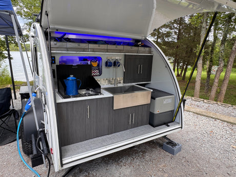Compact RV rental teardrop camper's illuminated outdoor kitchen with stove, sink, & fridge, set up by a lake.