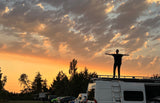 Experiencing golden hour from an RV rental! Person stands silhouetted on the roof, arms outstretched, admiring the colorful sky.