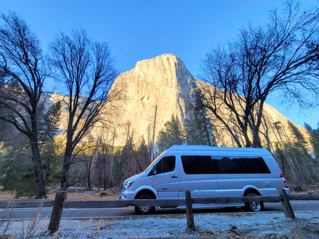 Light-colored RV rental in Yosemite Valley. Sunlit El Capitan rises behind bare trees and a wooden fence, under a clear sky.