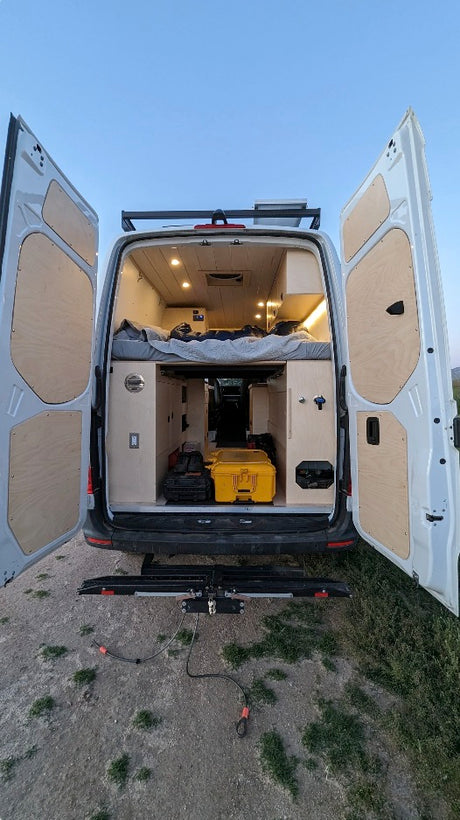 Experience mobile living with this RV rental! Open rear doors of a white camper van, showcasing a cozy bed, storage, and bike rack.