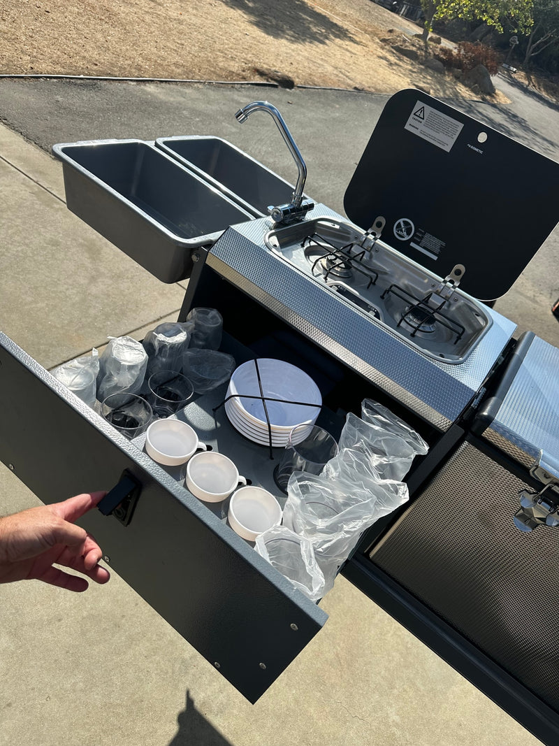 This portable RV rental kitchen features a sink, stove, and organized dinnerware, ready for outdoor cooking. — photo 12