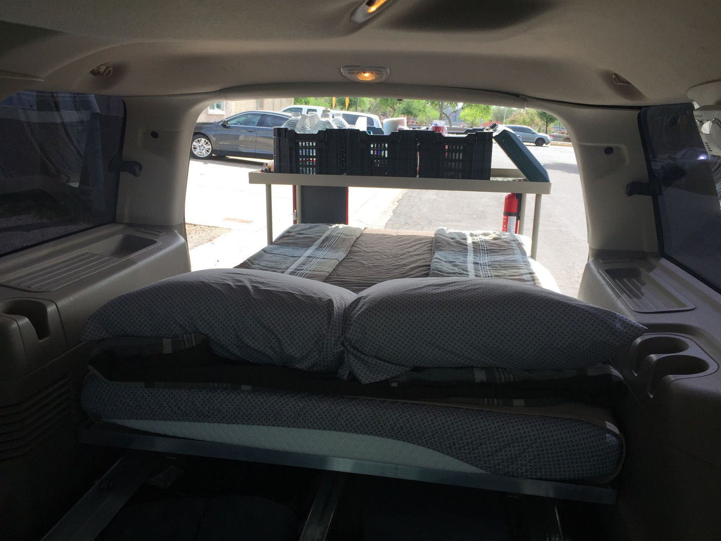 Inside a converted SUV 'rv rental': a cozy bed, pillows, and overhead storage crates for a comfortable car camping trip.