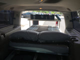 Inside a converted SUV 'rv rental': a cozy bed, pillows, and overhead storage crates for a comfortable car camping trip.