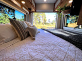 Inside a cozy RV rental: a comfy bed with decorative pillows & knit blanket overlooks a serene natural landscape.