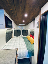Cozy RV rental interior: A built-in bed with colorful pillows, wooden ceiling, and compact kitchen features like a microwave.