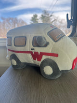 A cream plush RV toy with a red 'W' logo sits on a wood surface, against a blurred sky. Ready for your rv rental adventure!