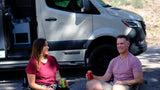 A smiling couple relaxes outside their Winnebago rv rental van with drinks, enjoying the outdoors on their trip.