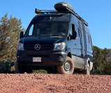 Black Mercedes Sprinter rv rental, equipped for adventure, parked on a reddish, rocky road under a clear sky.