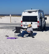 Relaxing at the beach! Person reads by their RV rental, with an empty chair, yoga mat, and ocean view.