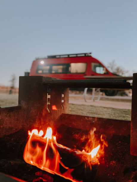 Bright campfire burns in a fire pit, with a red RV rental parked behind, under a fading sky.