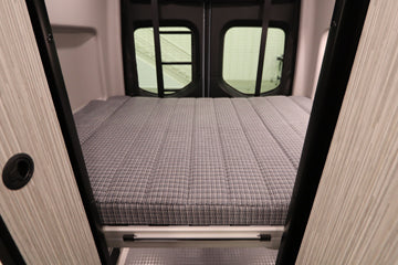Comfortable RV rental interior: A spacious, neatly made grey patterned bed is shown, ready for your road trip. Rear windows are visible.