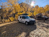 Sunny autumn day at a scenic campsite. White rv rental camper van amidst vibrant yellow fall foliage and fallen leaves.