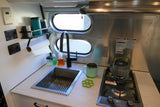 Compact kitchen in an RV rental, featuring a sink, two-burner stove, and monitoring displays for battery/water tanks.
