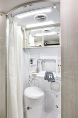 Compact RV rental wet bath. White shower curtain, toilet, sink, mirrored cabinet, and ceiling lights are visible.