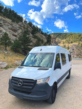 White RV rental van parked on a dirt road in scenic mountains under a bright blue sky with clouds.