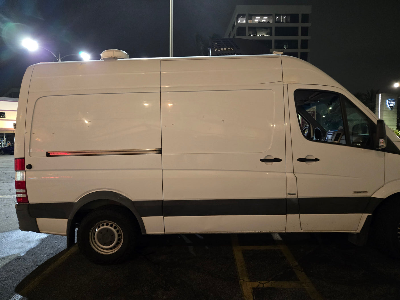 White Mercedes Sprinter camper van, possibly an RV rental, with a Furrion rooftop unit, parked at night.