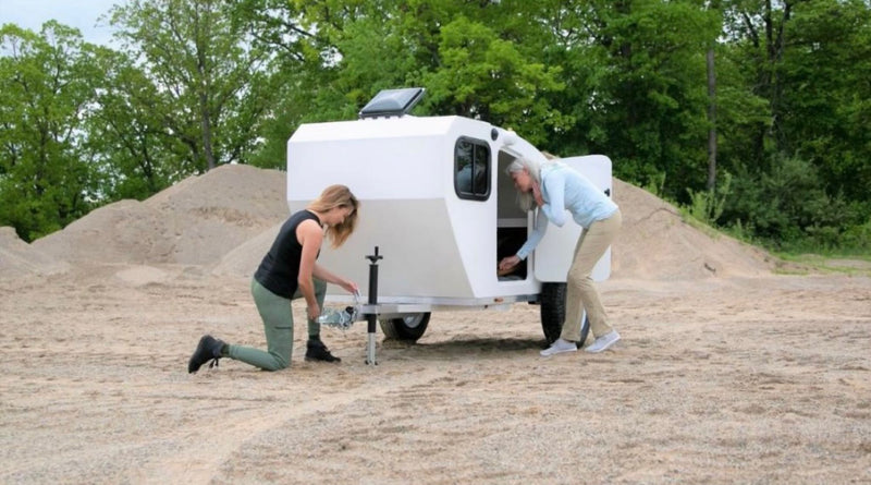  the other accesses the side storage of the compact camper.|A white teardrop RV rental hitched to a dark SUV on a dirt path with sand piles.|A woman sits in the open door of a white teardrop camper — photo 2