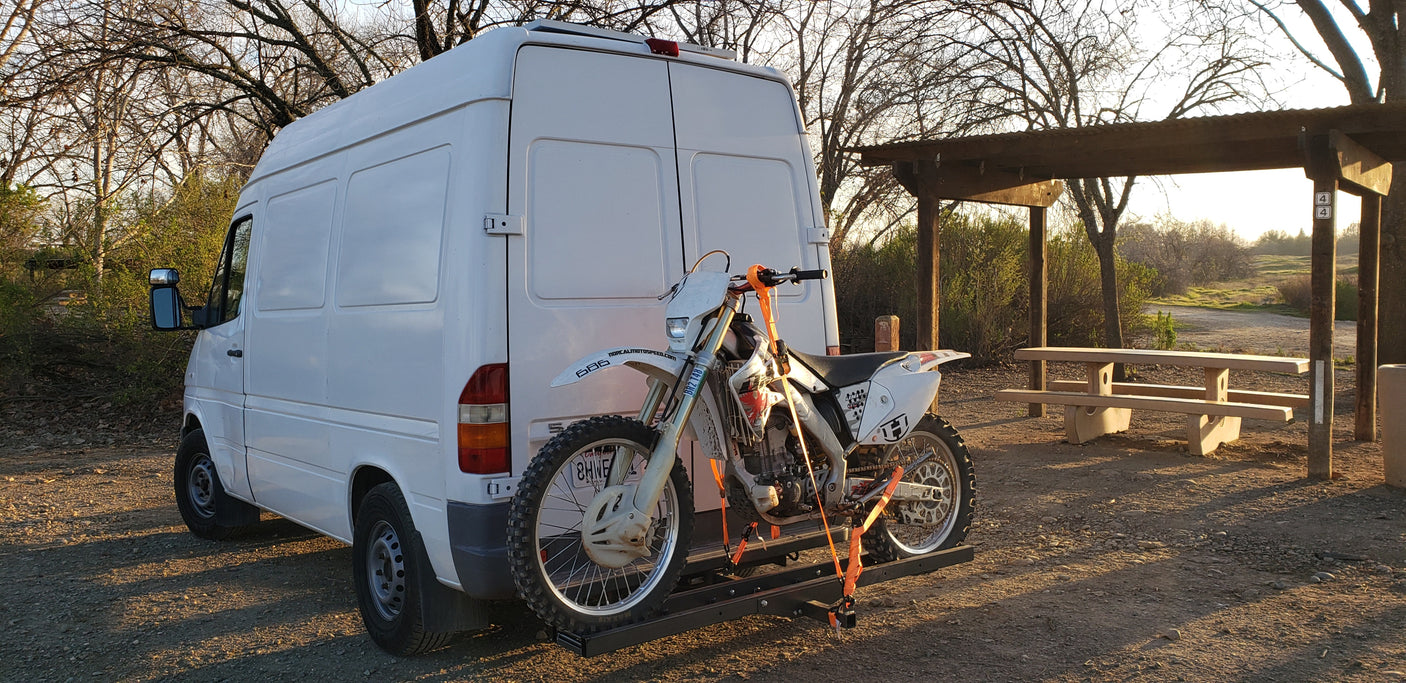 A white van carries a dirt bike on a rear rack, parked by a picnic area under bare trees. Great for outdoor adventures; consider an RV rental van.