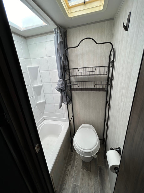RV rental bathroom: compact design with tub/shower, skylight, toilet, and metal storage shelf. Light walls and wood-look floor.
