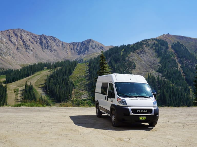White RV rental van parked on a dirt lot with scenic mountain peaks & green ski slopes under a clear blue sky.