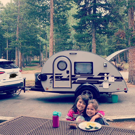 Happy girls at a picnic table, with a T@G teardrop RV rental & SUV parked in a scenic campground.
