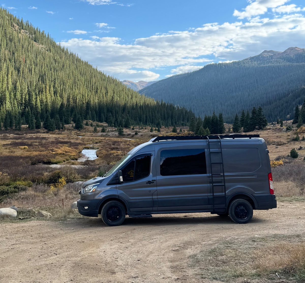 with a person warming by a fire pit under a lit awning at dusk in the mountains.|Gray RV rental camper van parked on a dirt road