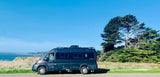 Dark grey Winnebago Travato RV rental parked on a coastal road overlooking ocean cliffs and green trees under a clear sky.