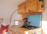 An RV rental's interior reveals a warm kitchen with a stove, sink, wood cabinets, and a nearby bed.