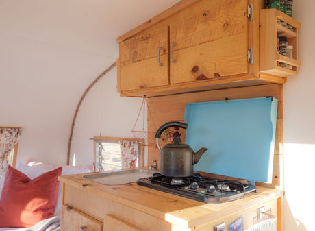 An RV rental's interior reveals a warm kitchen with a stove, sink, wood cabinets, and a nearby bed.