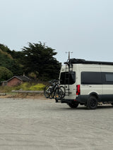 Winnebago Revel rv rental camper van with bikes & roof rack on a cloudy day, parked in a rustic, wooded area.