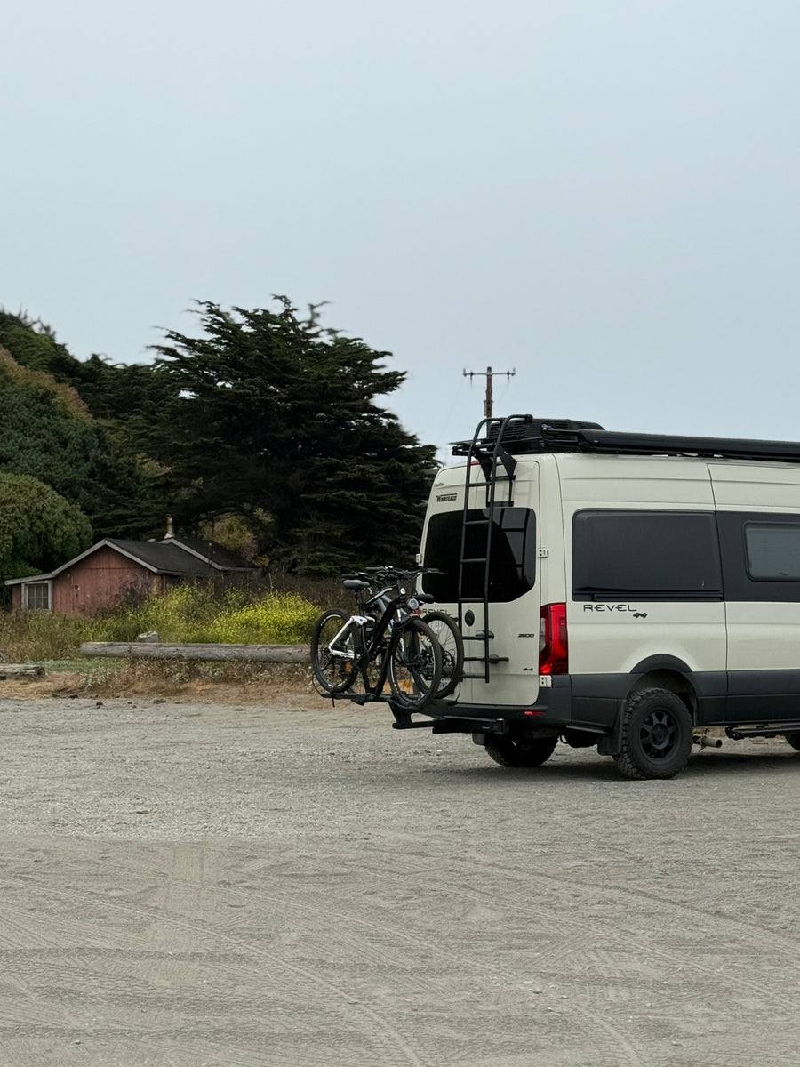 Winnebago Revel rv rental camper van with bikes & roof rack on a cloudy day, parked in a rustic, wooded area.