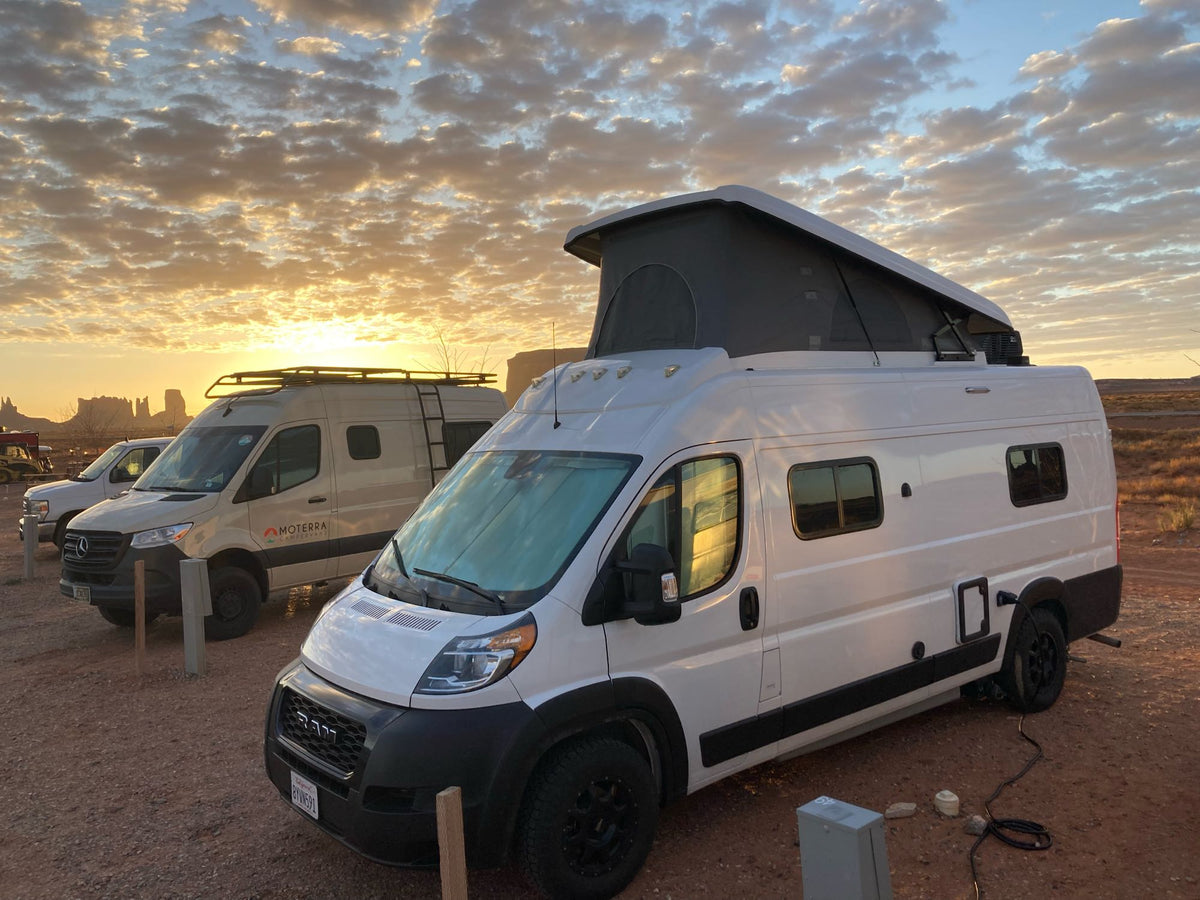 White pop-top campervan & tan RV rental parked in desert at stunning sunset with golden clouds. Epic van life adventure.|Relaxing at a wooded campsite with a pop-top RV rental