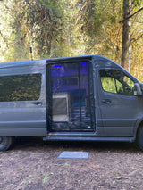 Open side of a gray RV rental camper van parked in a forest, revealing a bug screen & blue lit interior.