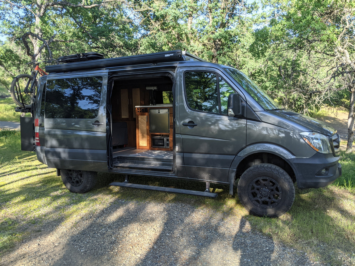 Explore in this rugged dark gray Sprinter RV rental camper van, featuring an open door revealing its interior, bike rack, & off-road tires.