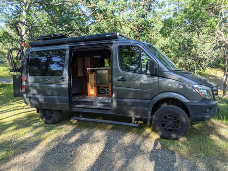 Explore in this rugged dark gray Sprinter RV rental camper van, featuring an open door revealing its interior, bike rack, & off-road tires.