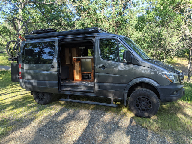 Explore in this rugged dark gray Sprinter RV rental camper van, featuring an open door revealing its interior, bike rack, & off-road tires.